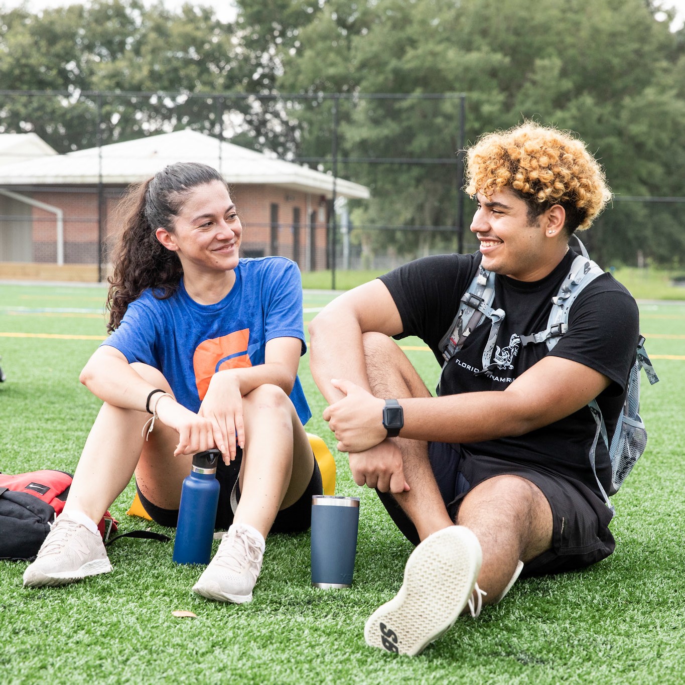 Two students sit on a field and chat.