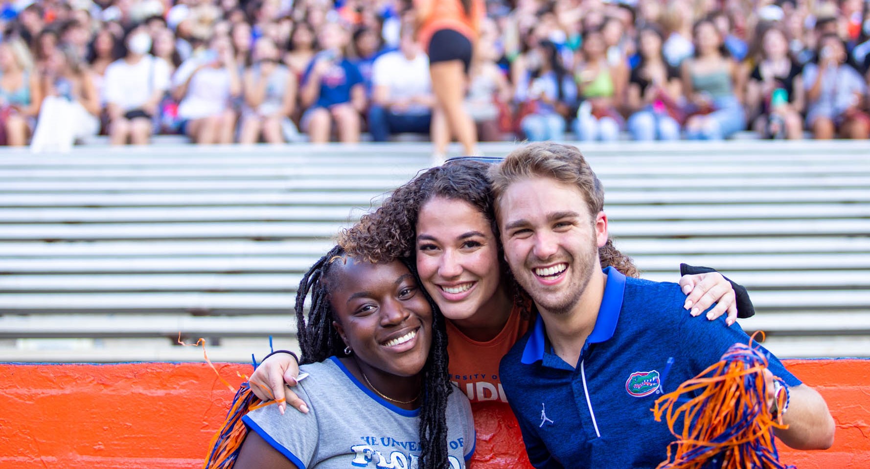 three students wear gator gear and smile in the football stadium.