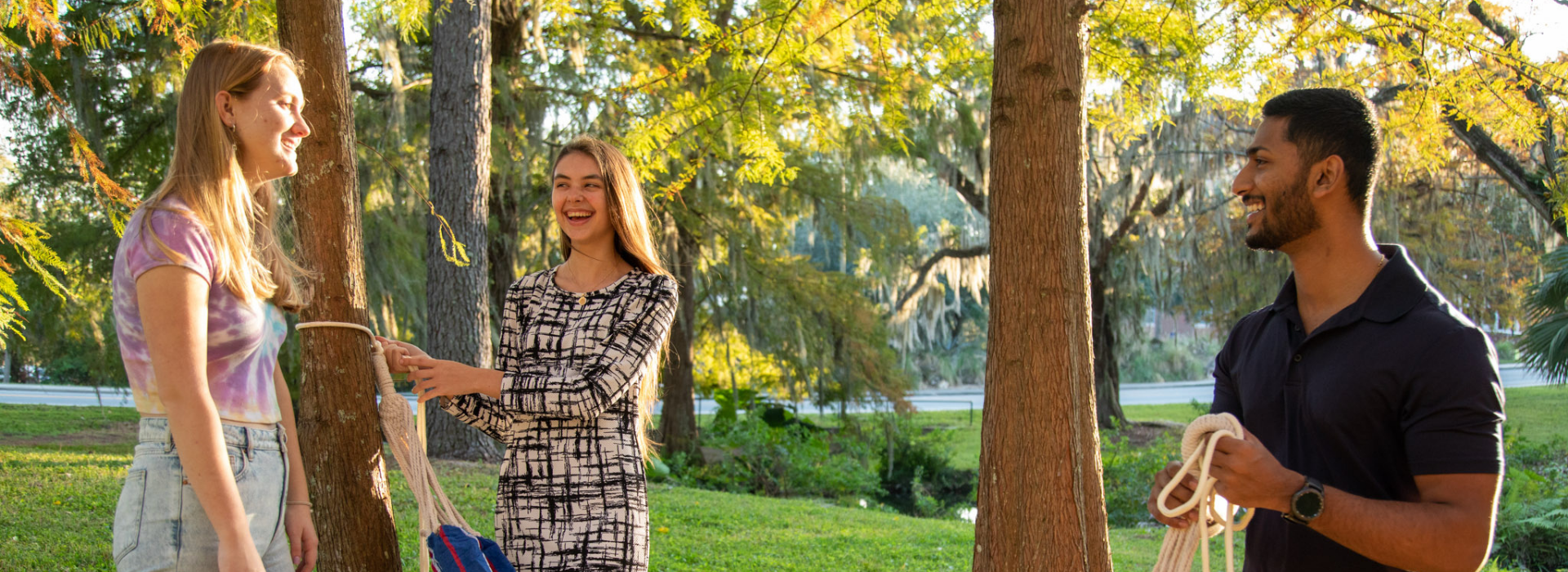 Three students hang a hammock between trees.