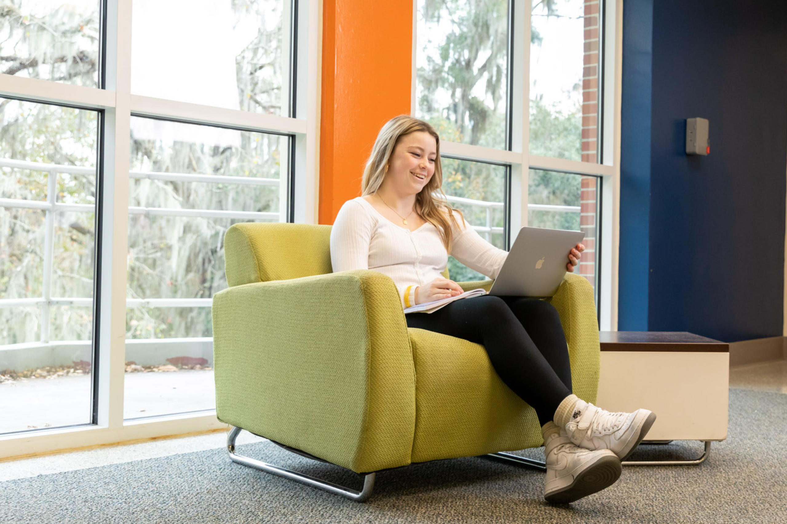 A blonde female student wears a white shirt, black leggings, and white sneakers while smiling and studying in her common room.