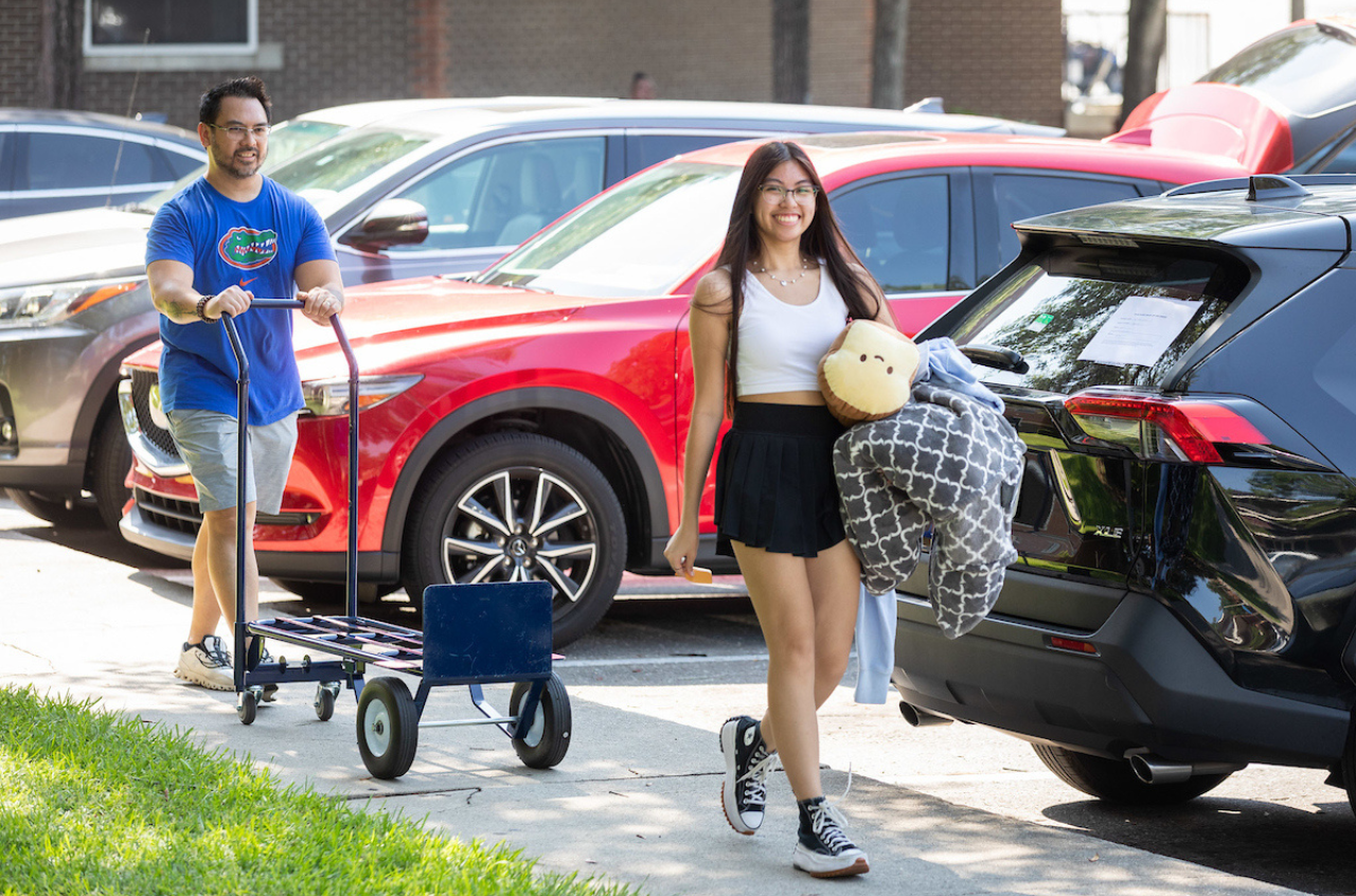 A student walks along the sidewalk of her hall during move-in. Her dad follows behind her with a cart.